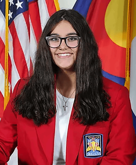 A girl wearing a red jacket and glasses sits in front of an American flag, showcasing a patriotic backdrop.