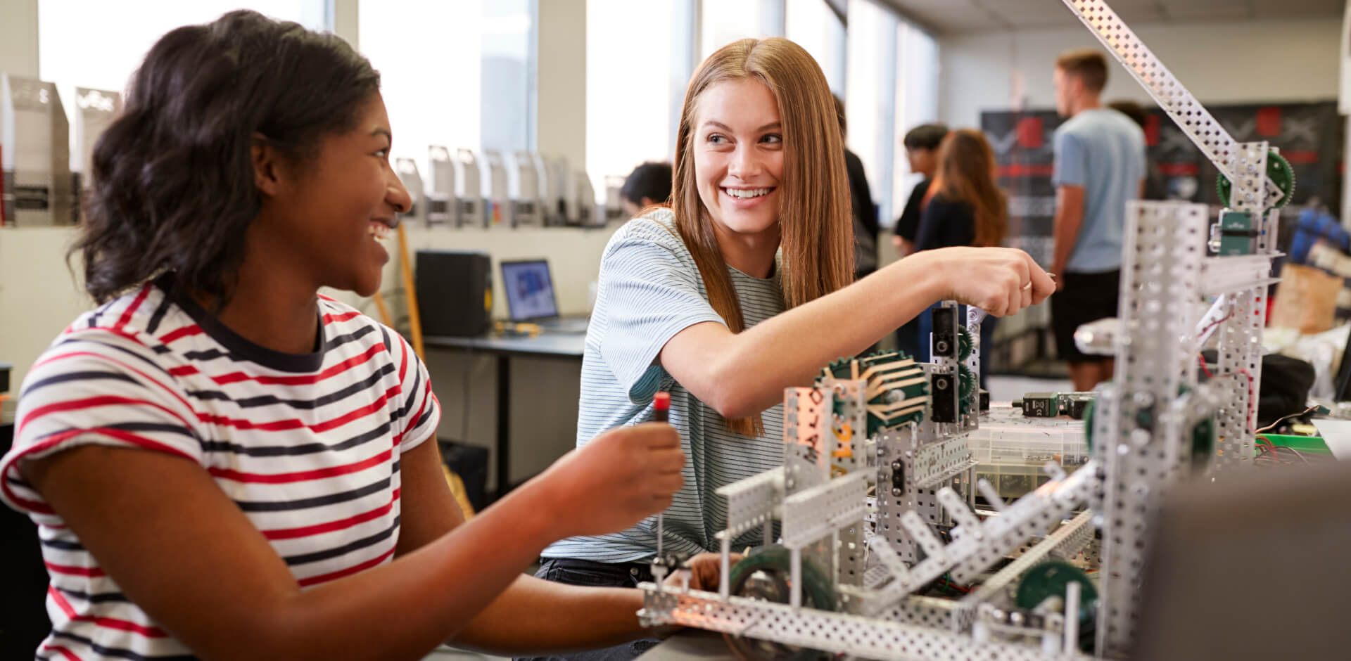 Two young women collaborate on building a robot in a classroom setting, showcasing teamwork and innovation in technology.