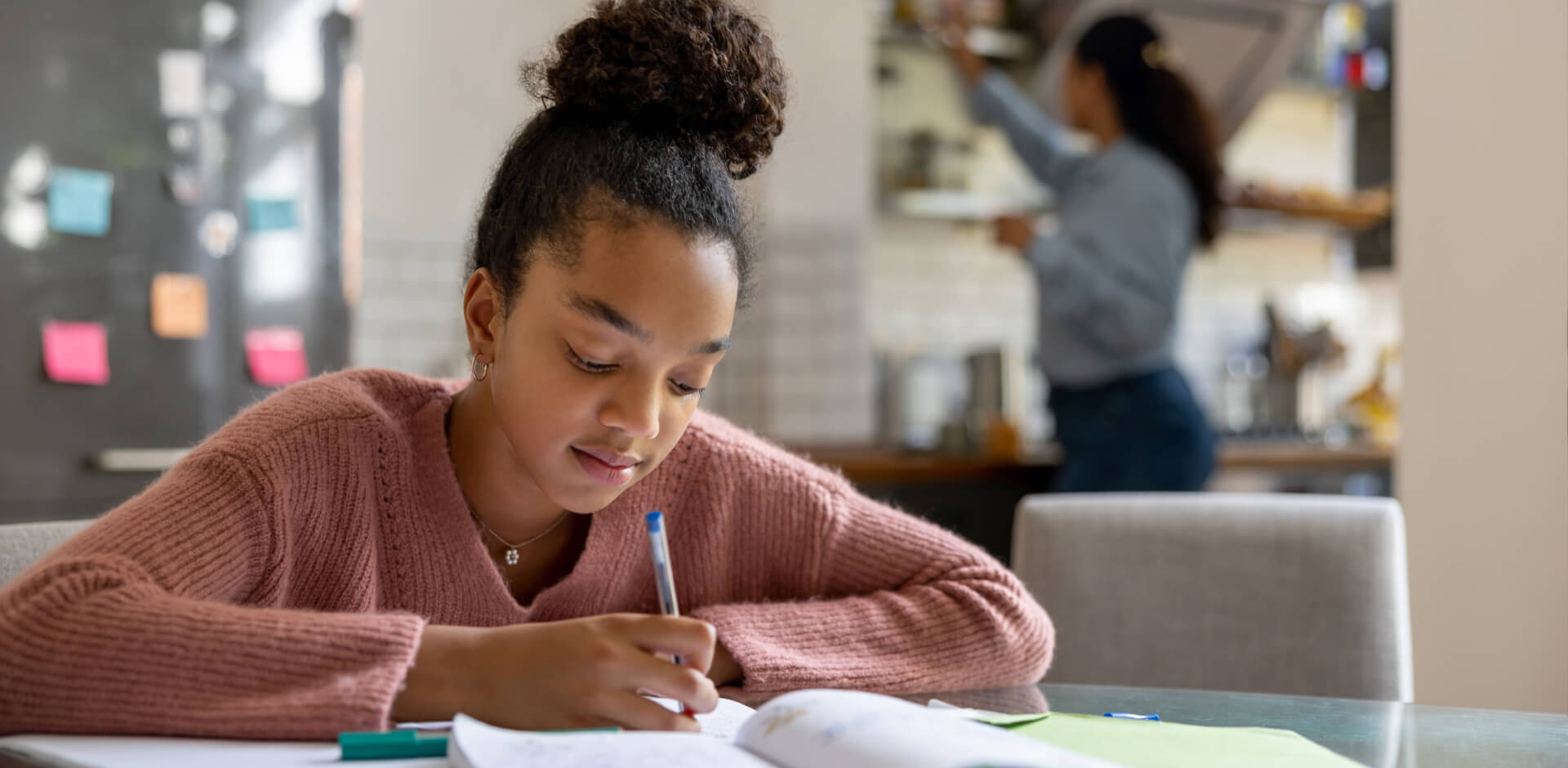 A young girl sits at a table, focused on writing in her notebook with a pencil, surrounded by colorful stationery.