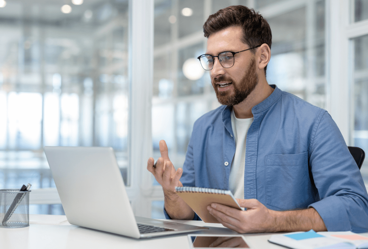 A man in glasses and a blue shirt sits at a desk in an office, engaged in a video call on a laptop, holding a notebook, appearing focused and friendly.