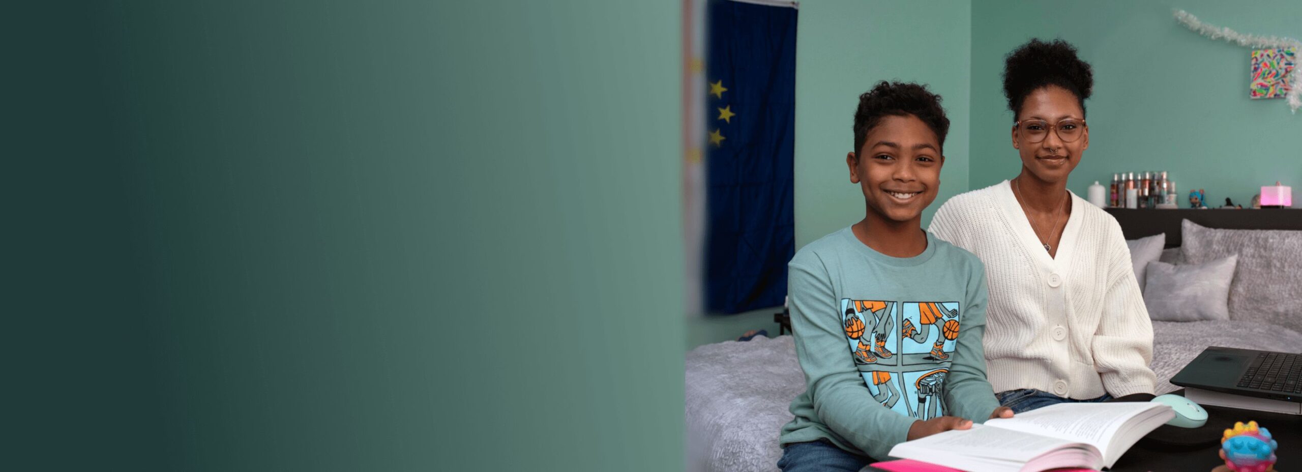 A young boy and a woman sit smiling at a table covered with books and a laptop. The room has a cozy vibe with soft colors and a flag on the wall.