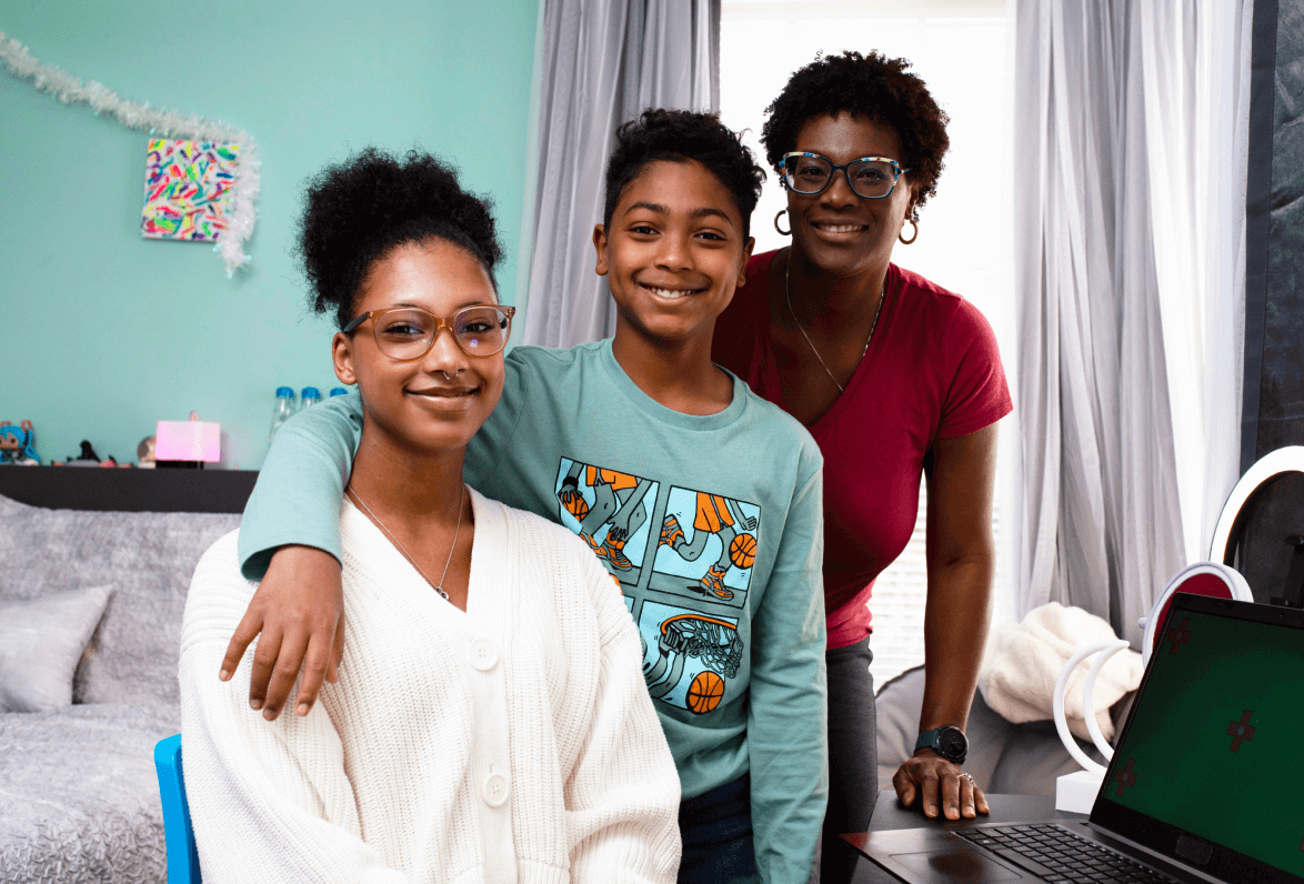 A smiling family of three, two children and an adult, pose together indoors. They are near a laptop, with a relaxed and cheerful atmosphere.