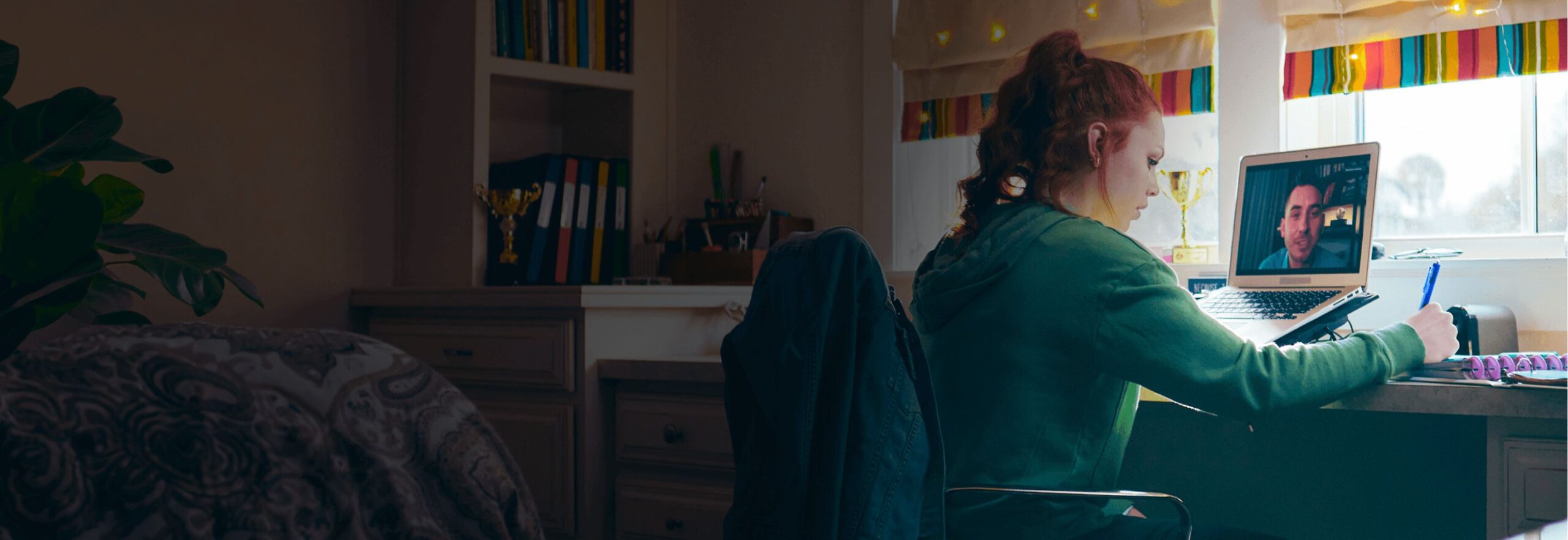 A young woman in a green hoodie writes notes at a desk while video-calling on a laptop. The room is cozy, with a plant and colorful blinds.