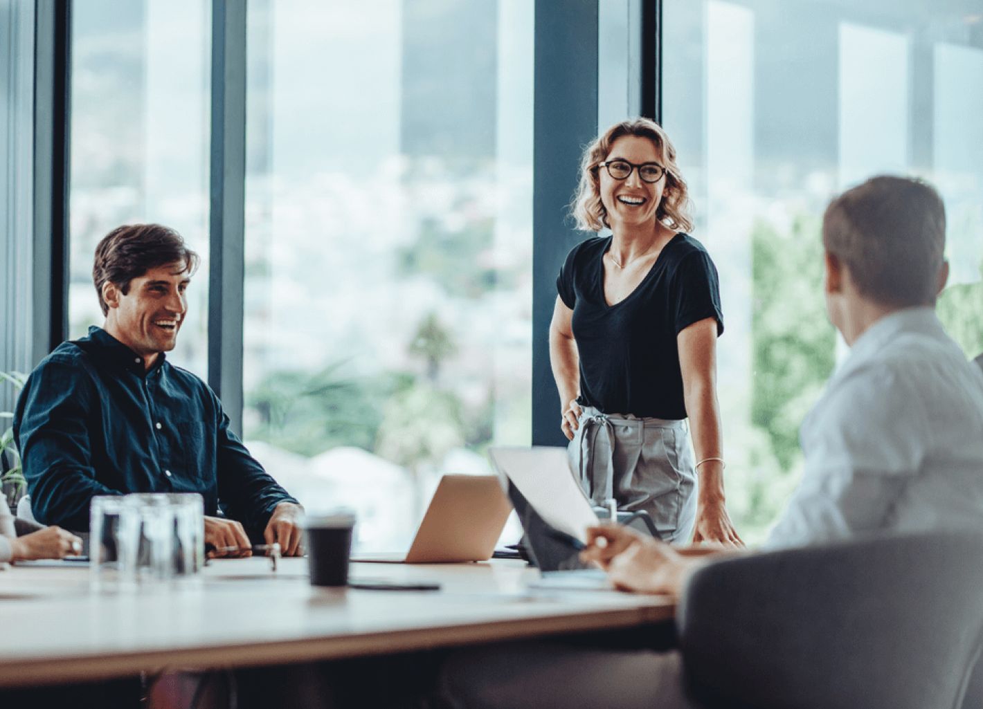 A group of four diverse colleagues laughing in a bright office. A woman stands confidently, engaging the team. The mood is positive and collaborative.