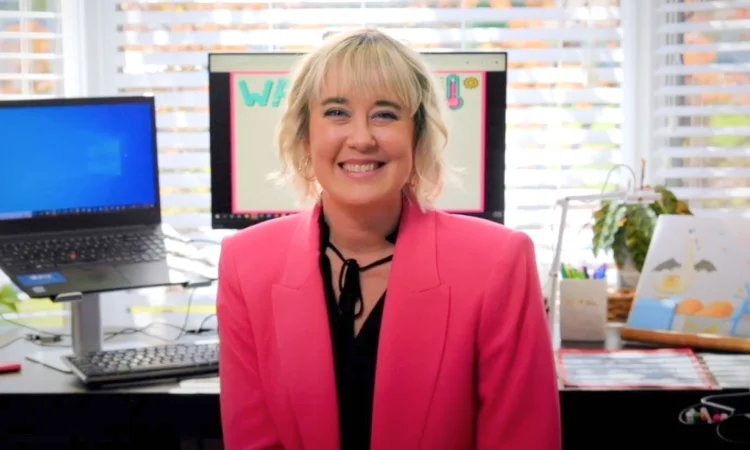 A person in a pink blazer smiles warmly at a desk with open laptops and cheerful decor. The setting conveys a welcoming and positive vibe.