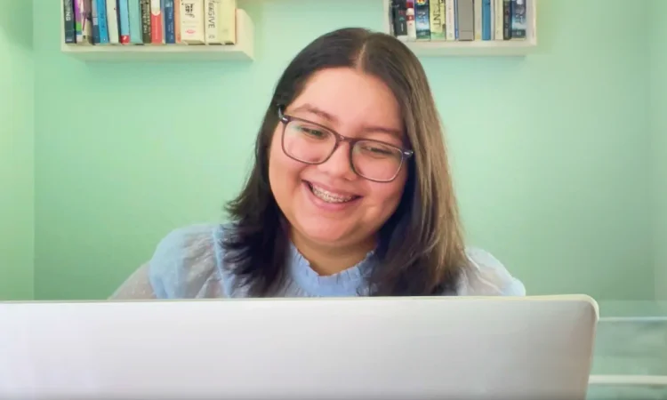 Smiling person with braces and glasses looks at a laptop. Background has mint green walls with books on shelves, creating a cozy, focused atmosphere.