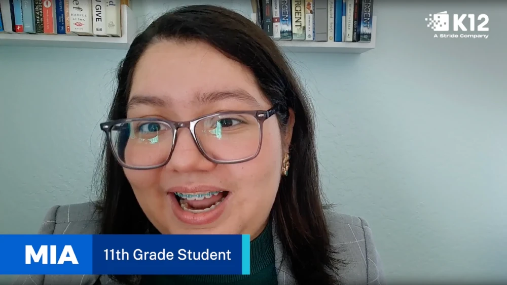 Student speaking during a webcam-style video interview from a home study space, with a bookshelf in the background and a lower-third label reading 'MIA, 11th Grade Student.