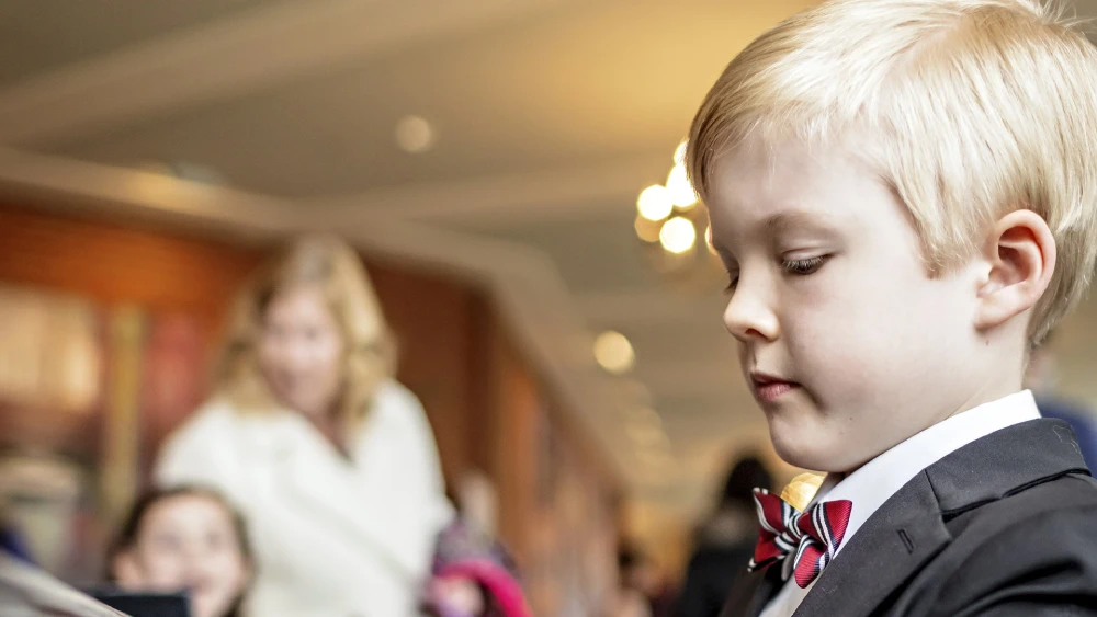Close-up of a young student in formal attire reading or looking at learning materials in a classroom setting, with other people blurred in the background.